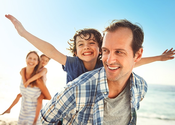 A joyful father giving his son a piggyback ride on a sunny beach.